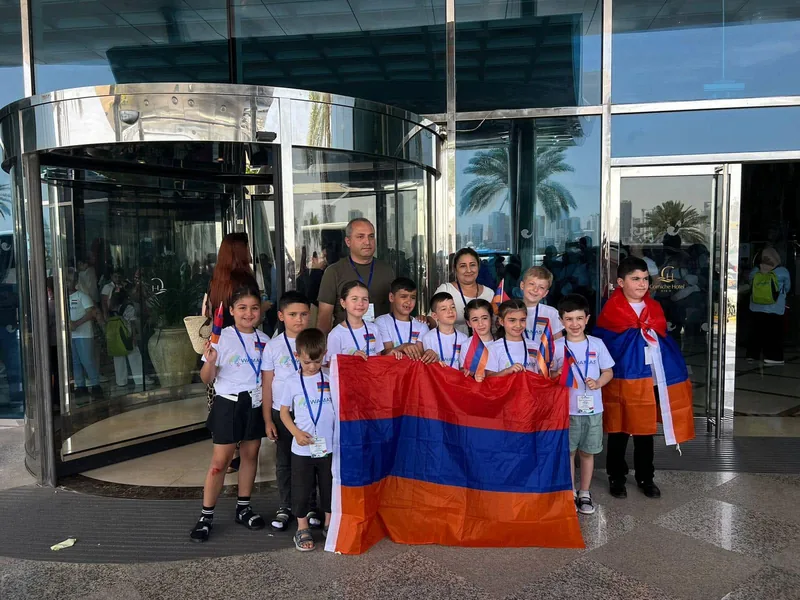 WAMAS-ի շապիկներով երեխաներն ու մեծահասակները Հայաստանի դրոշներով՝ հյուրանոցի ապակե մուտքի մոտ, մենթալ թվաբանության մրցույթի ուղևորության ընթացքում։