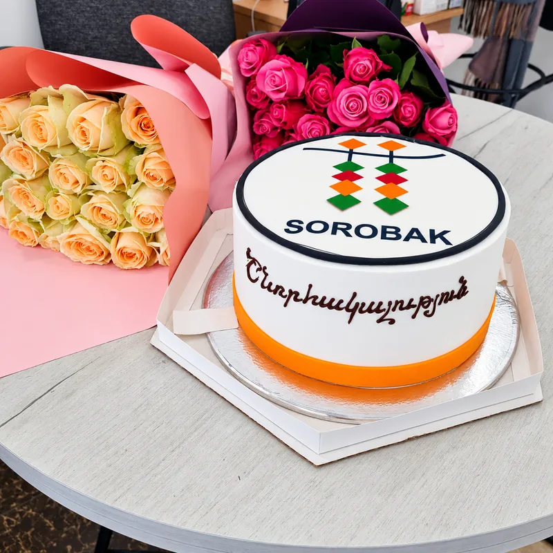 Sorobak-ի տարբերանշանով սպիտակ տորթ՝ նարնջագույն եզրով, դեղձագույն և վարդագույն վարդերի ծաղկեփնջերի կողքին։